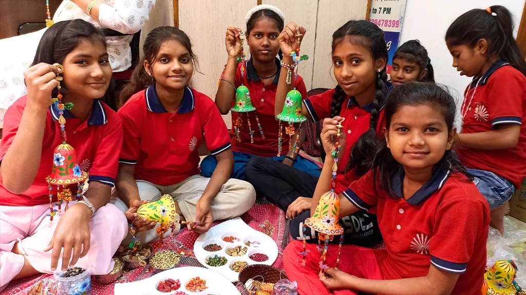 Smiling girls in red uniforms at Divine Sanskarshala holding handmade colorful crafts during an arts and crafts session.