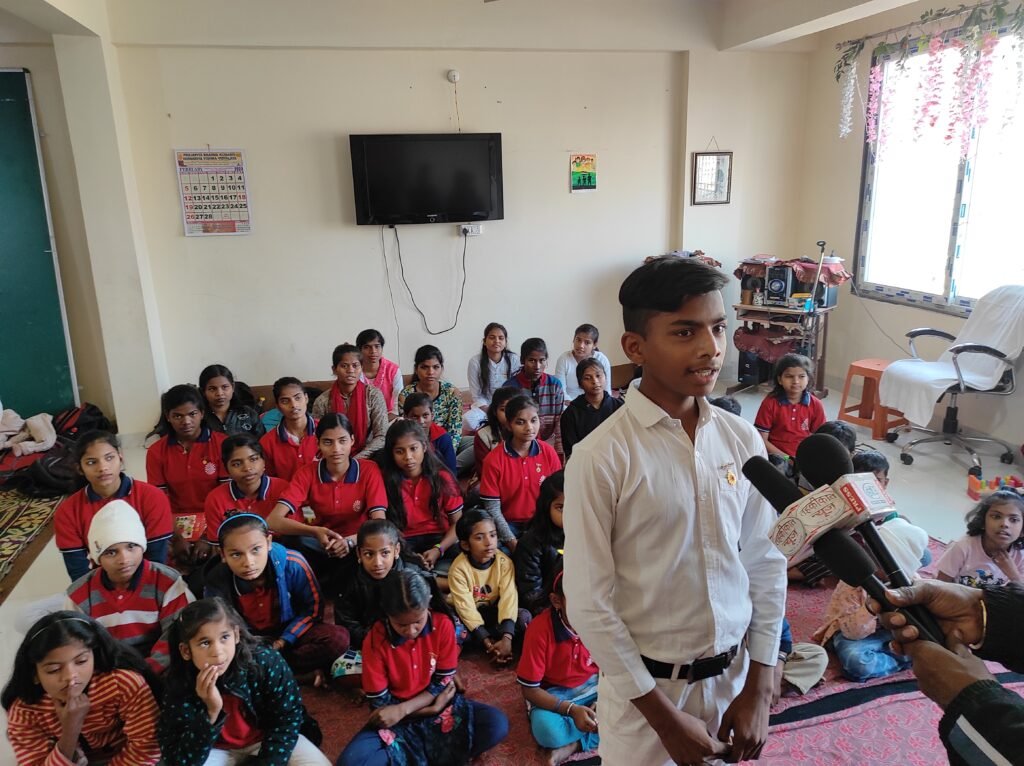 A boy in white uniform speaking confidently into microphones while other children of Divine Sanskarshala sit attentively behind him during a media interaction.