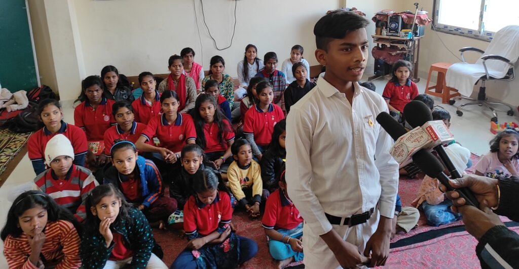 A boy in white uniform speaking confidently into microphones while other children of Divine Sanskarshala sit attentively behind him during a media interaction.
