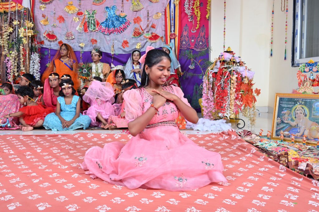 Young girl performing a traditional dance on stage while other children in colorful cultural costumes sit behind during a school cultural program.