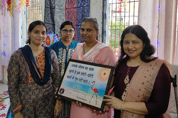 Four women stand together indoors during a recognition ceremony at Divine Sanskarshala. One woman in the center holds a framed appreciation certificate while the others stand beside her smiling. The room is decorated with colorful hanging flowers, lights, and patterned curtains.