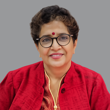 Portrait of Kalpana Gupta, Executive Member of Divine Sanskarshala, smiling and wearing glasses and a red traditional jacket against a gray background.