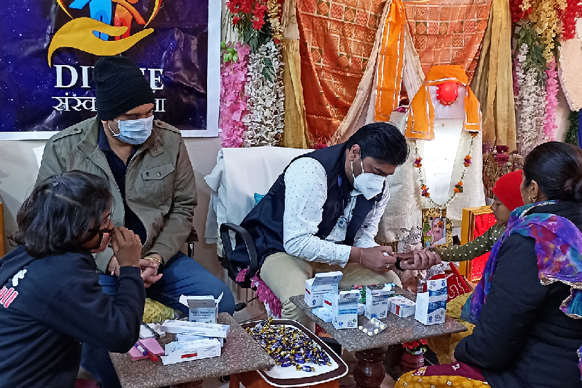 Healthcare workers wearing face masks provide medical consultation and medicines to women and children during a Divine Sanskarshala community health camp in a decorated indoor setting.