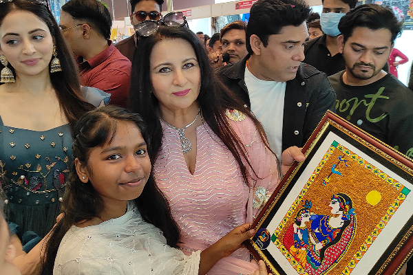 Actress Poonam Dhillon posing with a student from Divine Sanskarshala who is presenting a colorful traditional artwork at a public event surrounded by attendees.