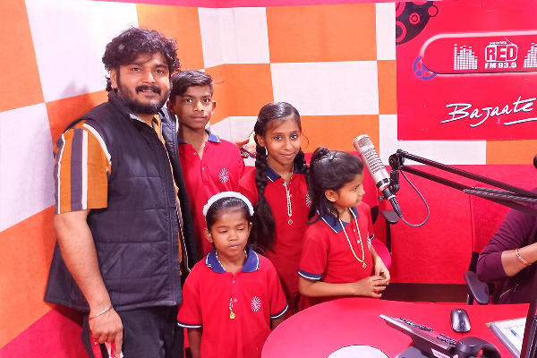 RJ Shashank stands with a group of children from Divine Sanskarshala inside a Red FM radio studio. The children are wearing matching red school uniforms and standing near a microphone and broadcasting desk.