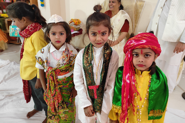 Three young children dressed in colorful traditional Indian outfits stand together during a cultural celebration at Divine Sanskarshala. They wear vibrant scarves, festive accessories, and traditional makeup, while other participants and decorations appear in the background.