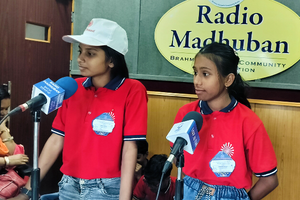 Two girls wearing red Divine Sanskarshala uniforms stand in front of microphones inside the Radio Madhuban studio. They appear to be speaking or participating in a radio program while people sit in the background.