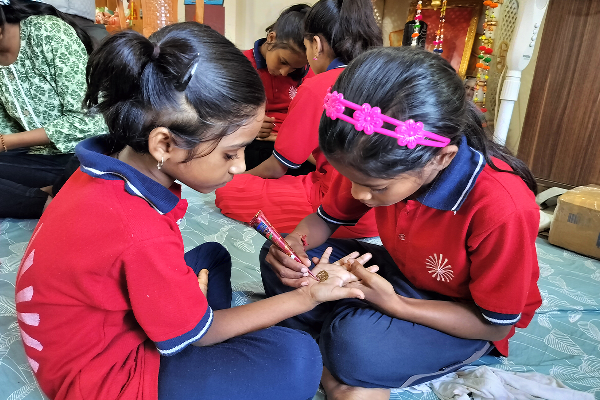 Two young girls wearing red Divine Sanskarshala uniforms sit together while one carefully applies mehndi (henna) on the other’s hand during a creative activity session. Other students can be seen participating in activities in the background.