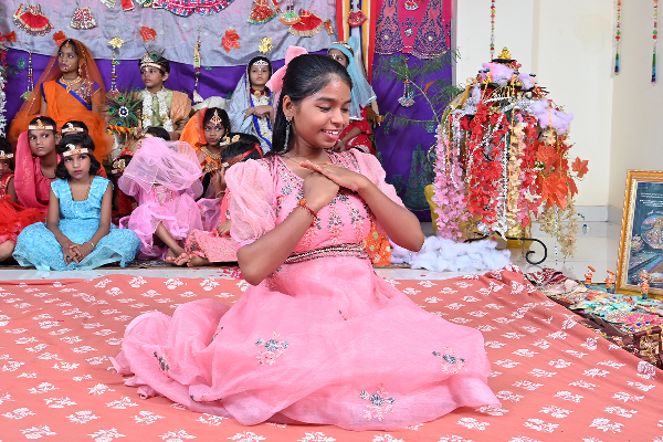A young girl in a pink traditional dress performs a graceful dance at a cultural event organized by Divine Sanskarshala. Several children in colorful traditional attire are seated in the decorated background, surrounded by vibrant handmade decorations and festive artwork.