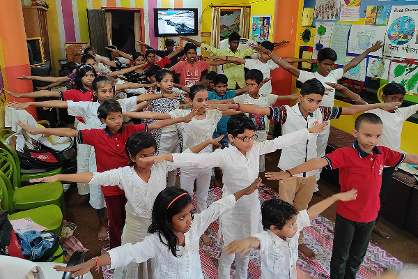 A large group of children at Divine Sanskarshala participate in a group exercise session indoors, standing with their arms stretched out in unison. The colorful classroom is decorated with educational charts, artwork, and bright walls.