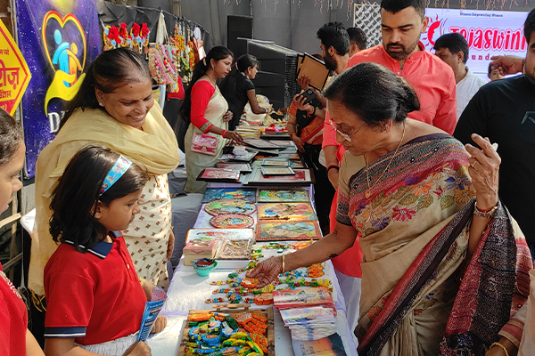 Rita Bahuguna Joshi visits the Divine Sanskarshala stall, interacting with children and observing handmade educational and artistic creations displayed on tables during an exhibition.