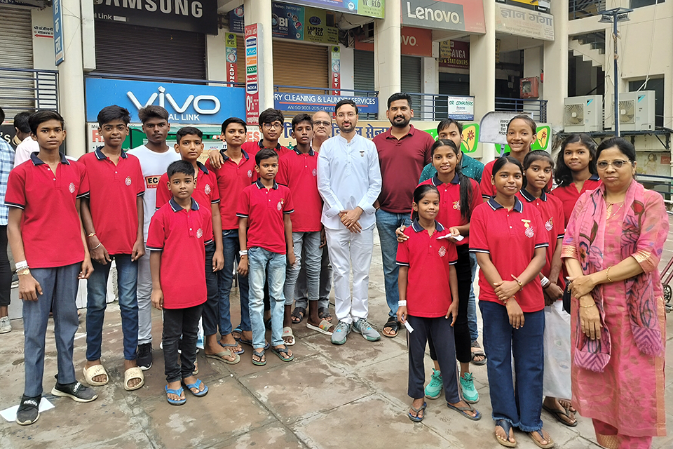 Group photo of Divine Sanskarshala children, volunteers, and organizers standing together during a community cyclothon awareness event. The children are wearing matching red uniforms and smiling alongside adult supporters in an outdoor public area.