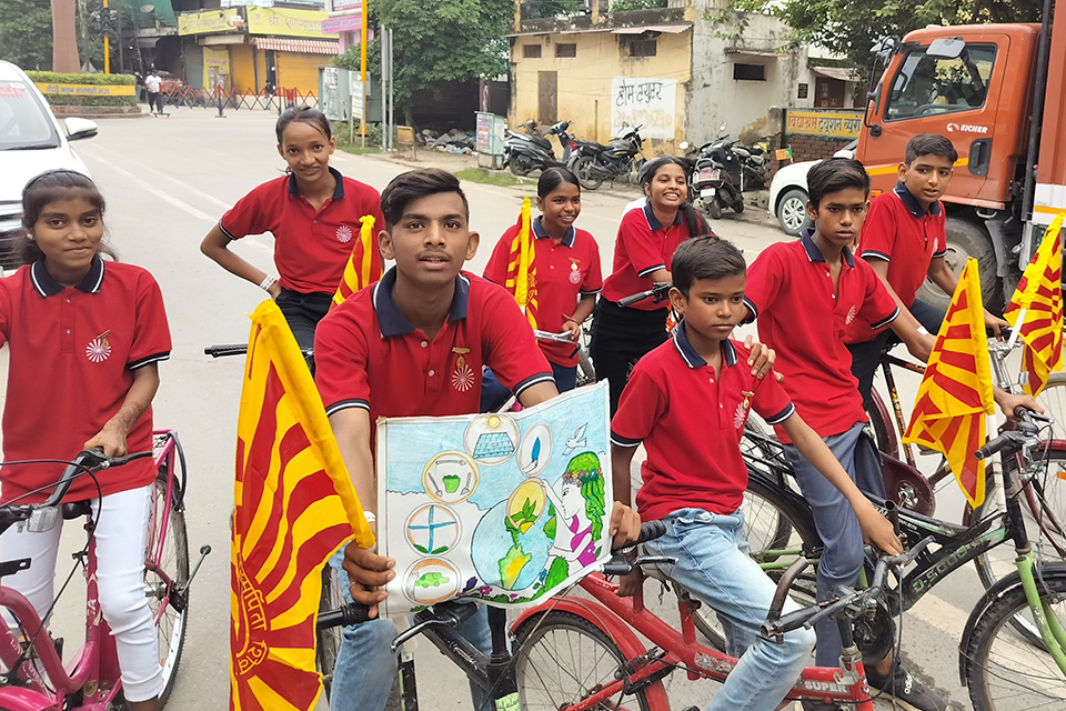 Children of Divine Sanskarshala participate in a cyclothon, riding bicycles with awareness flags and posters promoting environmental conservation and healthy living on a city road.