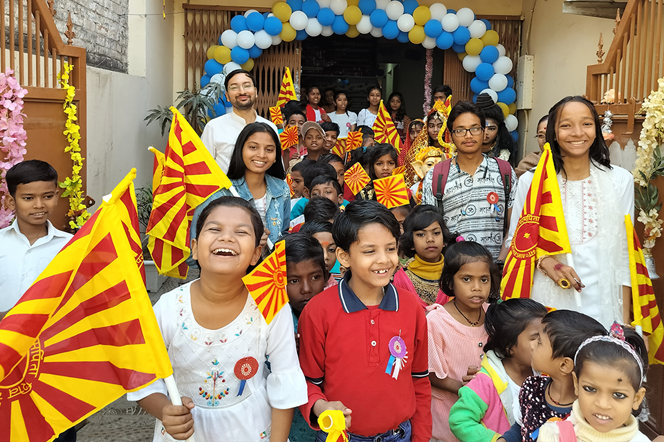 Children and volunteers from Divine Sanskarshala holding colorful flags and smiling together during a Shiv Jayanti celebration, gathered at the entrance of a decorated venue with balloon arches.