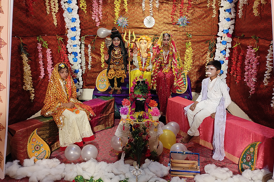 Children dressed as Lord Shiva, Goddess Parvati, Lord Ganesha, and other mythological characters perform during a Maha Shivratri celebration at Divine Sanskarshala. The stage is beautifully decorated with flowers, balloons, and traditional festive décor.