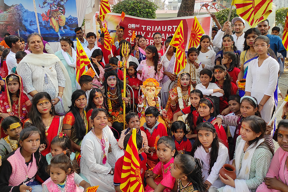 Children, volunteers, and supporters of Divine Sanskarshala participate in the 82nd Trimurti Shiv Jayanti celebration, holding saffron flags and posing with children dressed as Lord Shiva, Goddess Parvati, and Lord Ganesha during a cultural procession.