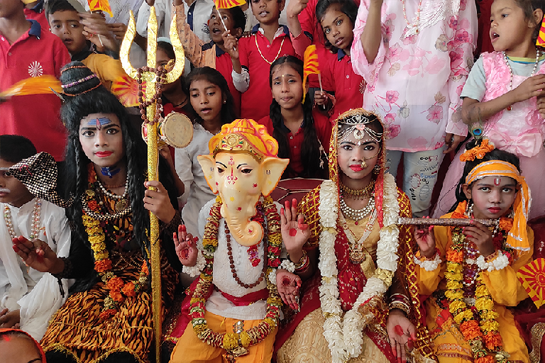 Children dressed as Lord Shiva, Lord Ganesha, Goddess Parvati, and Lord Krishna participate in a Maha Shivratri cultural performance at Divine Sanskarshala, surrounded by fellow students holding saffron flags and celebrating with devotion.