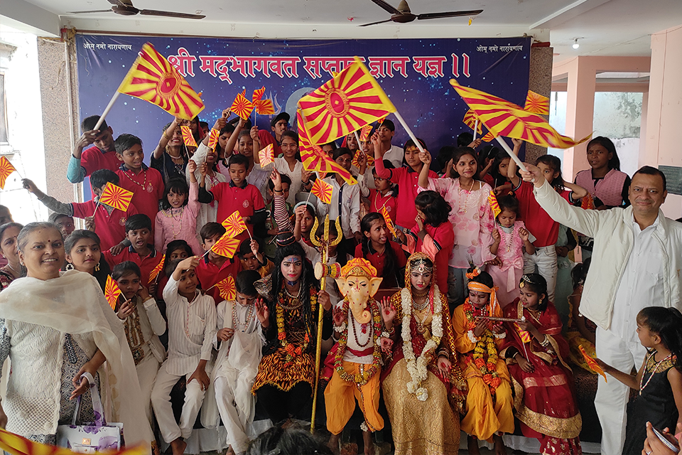 Children of Divine Sanskarshala, dressed as Lord Shiva, Lord Ganesha, Goddess Parvati, and Lord Krishna, celebrate Maha Shivratri with teachers, volunteers, and supporters while waving saffron flags during a cultural gathering.
