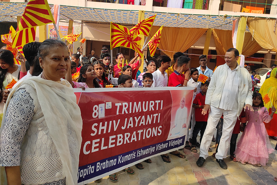 Children, volunteers, and community members from Divine Sanskarshala participate in the 81st Trimurti Shiv Jayanti celebrations, holding colorful flags and a large event banner during a festive procession.