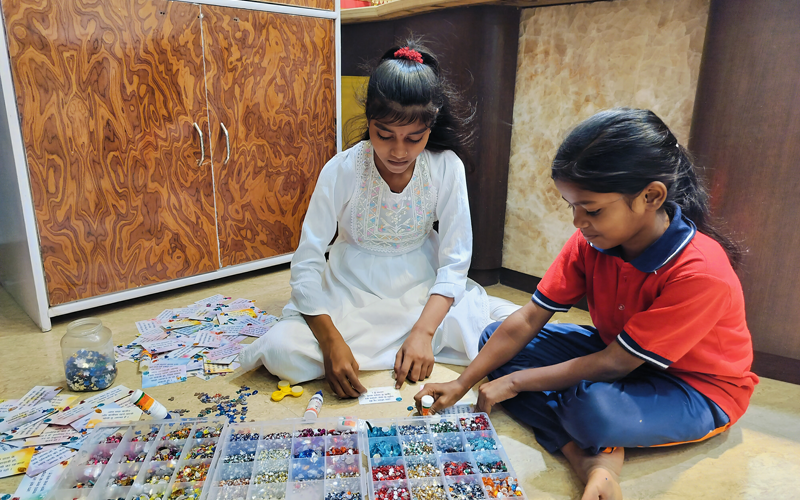 Two young girls at Divine Sanskarshala carefully making handmade beaded crafts using colorful beads and craft materials while sitting on the floor in a creative learning environment.